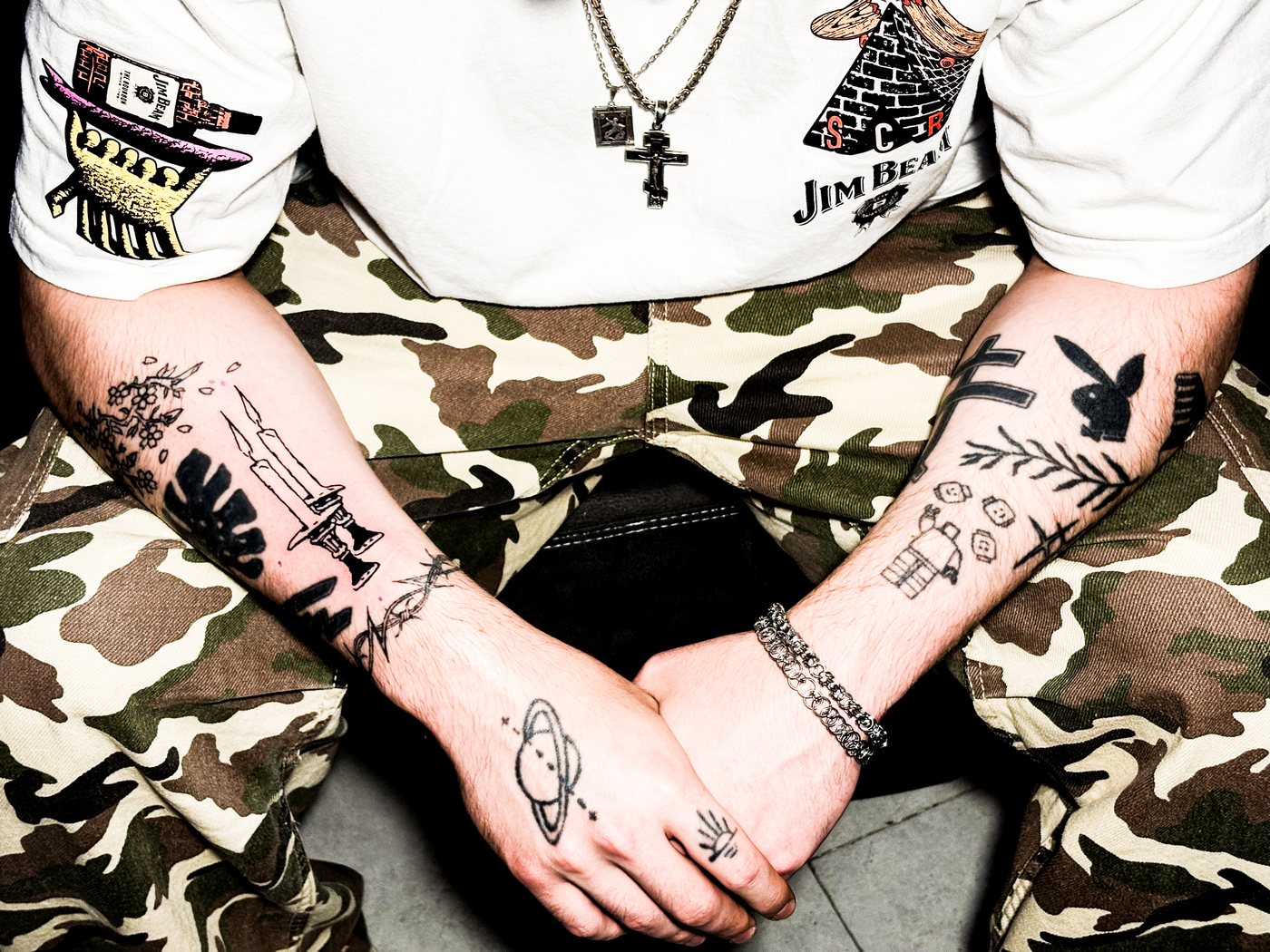 fine line patchwork tattoos across both forearms and hands, camo pants